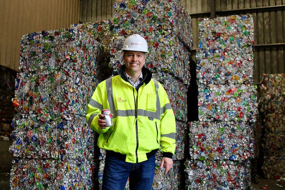 A Dem-Con employee poses for the camera with a can in hand. Behind him are bales of crushed cans